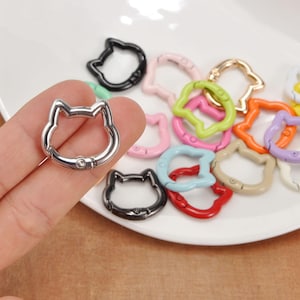 Puede incluir: Una colecci&oacute;n de cierres met&aacute;licos con forma de gato en varios colores, incluyendo plata, negro, rosa, verde, naranja y morado. Los cierres est&aacute;n dispuestos sobre un plato blanco, con un cierre plateado en una mano.