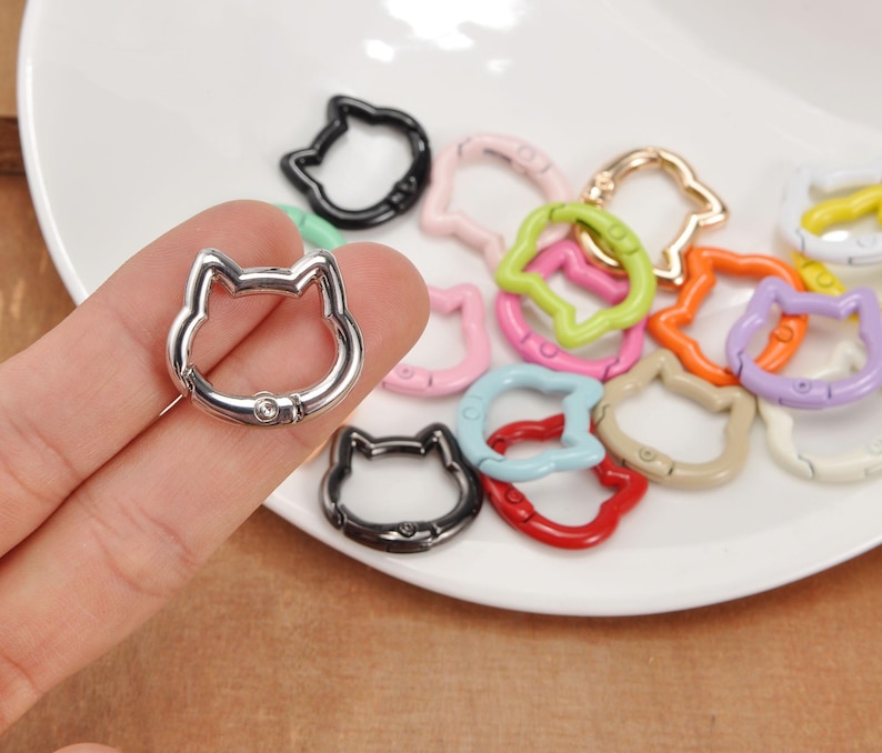 Puede incluir: Una colecci&oacute;n de cierres met&aacute;licos con forma de gato en varios colores, incluyendo plata, negro, rosa, verde, naranja y morado. Los cierres est&aacute;n dispuestos sobre un plato blanco, con un cierre plateado en una mano.