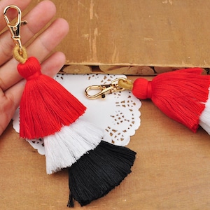 May include: A red, white, and black tassel keychain with a gold metal clasp. The tassel is made of three layers of fabric, with the top layer being red, the middle layer being white, and the bottom layer being black.