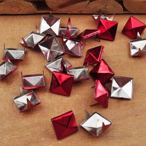 Puede incluir: Una colecci&oacute;n de tachuelas met&aacute;licas en forma de pir&aacute;mide, en plata y rojo. Las tachuelas tienen una base puntiaguda y est&aacute;n dise&ntilde;adas para sujetarse a tela o cuero.
