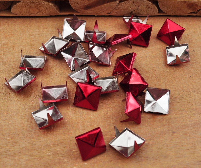 Puede incluir: Una colecci&oacute;n de tachuelas met&aacute;licas en forma de pir&aacute;mide, en plata y rojo. Las tachuelas tienen una base puntiaguda y est&aacute;n dise&ntilde;adas para sujetarse a tela o cuero.