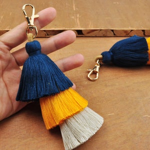 May include: A layered tassel keychain with a gold-colored clasp. The tassel features three tiers of color: navy blue, yellow, and off-white. The keychain is designed for attaching to keys or bags.