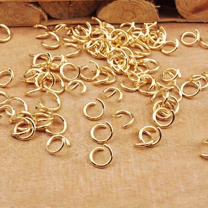 May include: A large quantity of gold-colored jump rings, scattered on a wooden surface. The rings are small and circular, with a gap in the circle.