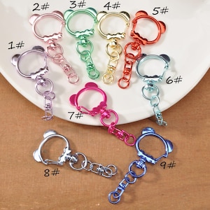 May include: A collection of nine metal keychains in various colors, including silver, pink, green, gold, red, and blue. Each keychain features a bear-shaped design with a clasp and chain. The keychains are arranged on a white plate and a brown surface.