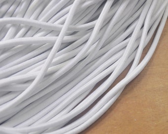 May include: Close-up of a pile of white elastic cord. The cords are tightly packed together, creating a dense, textured surface. The cords are smooth and cylindrical, with a slight sheen. The background is a light brown wooden surface.