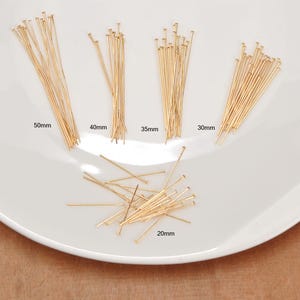 Puede incluir: Un conjunto de alfileres de cabeza dorados de varias longitudes, incluyendo 50 mm, 40 mm, 35 mm, 30 mm y 20 mm. Los alfileres están dispuestos sobre un plato blanco, mostrando sus diferentes tamaños. Estos alfileres son ideales para la creación de joyas y manualidades.
