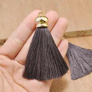 May include: Two dark gray silk tassels with gold metal tops. The tassels are held in a hand.