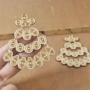 May include: Two wooden ornaments shaped like a stylised tree with a repeating eye pattern. The ornaments are made of light brown wood and have a delicate, intricate design.