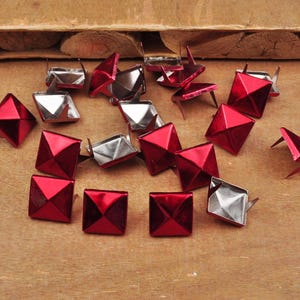Puede incluir: Una colecci&oacute;n de tachuelas de metal rojo en forma de pir&aacute;mide con bases plateadas. Las tachuelas est&aacute;n esparcidas sobre una superficie de madera.