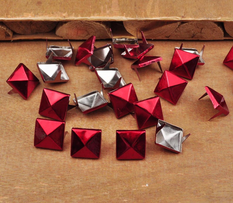 Puede incluir: Una colecci&oacute;n de tachuelas de metal rojo en forma de pir&aacute;mide con bases plateadas. Las tachuelas est&aacute;n esparcidas sobre una superficie de madera.