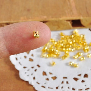 May include: A tiny gold-colored charm shaped like a bunny, resting on a fingertip. The charm has a small loop at the top. A scattering of similar charms is visible in the background, suggesting a jewelry-making theme.