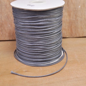May include: A spool of gray waxed cotton cord. The cord is tightly wound around a white cardboard core. The end of the cord extends from the spool, ready for use. The background is a light brown wooden surface.