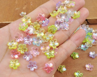 May include: Assortment of iridescent plastic flower-shaped beads in shades of pink, green, and yellow. The beads are scattered on a hand and a wooden surface.