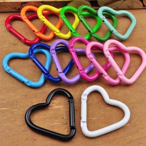 May include: A collection of heart-shaped carabiner clips in various colors, including red, orange, yellow, green, blue, purple, pink, black, and white. These metal clips are designed for attaching items and are arranged on a wooden surface.