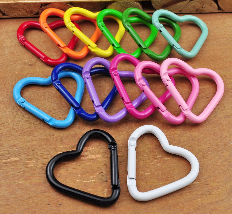 May include: A collection of heart-shaped carabiner clips in various colors, including red, orange, yellow, green, blue, purple, pink, black, and white. These metal clips are designed for attaching items and are arranged on a wooden surface.
