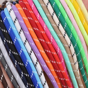 May include: A collection of multi-coloured paracord ropes arranged side by side. Each cord features a woven texture with a repeating white or black diagonal dashed pattern. The colours include brown, grey, black, blue, light blue, white, purple, orange, red, teal, beige, green, light green, pink, and yellow.