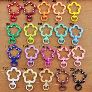 May include: Twenty flower-shaped metal keychains with different colored enamel paint and designs. Each keychain has a small metal ring attached to the top.
