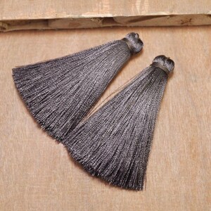 May include: Two gray silk tassels with a long, thin fringe. The tassels are on a light brown wooden surface.