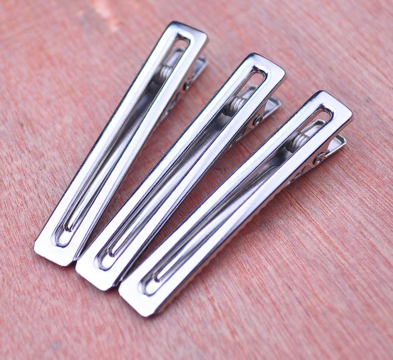 Op de afbeelding: Drie glanzende, zilverkleurige metalen haarclips diagonaal gerangschikt op een lichtbruin houten oppervlak. Elke clip heeft een rechthoekige vorm met een veermechanisme. De clips zijn reflecterend.