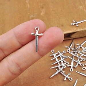 May include: Silver sword charms, a single charm is held in a hand, and a group of charms are scattered on a wooden surface.