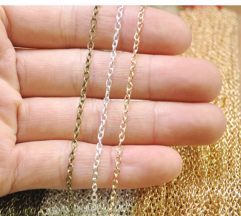 Puede incluir: Tres hebras de cadena en colores dorado, plateado y bronce. Las cadenas se sostienen en una mano, y el fondo es una pila de cadenas similares.