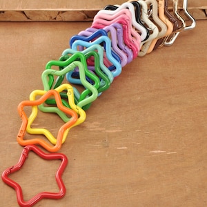 Puede incluir: Una colección de cierres metálicos en forma de estrella de varios colores, incluyendo rojo, naranja, amarillo, verde, azul, rosa y marrón. Los cierres están dispuestos diagonalmente sobre una superficie de madera, mostrando sus vibrantes colores y diseño.