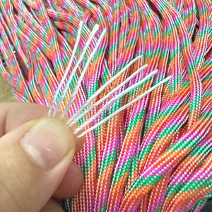May include: Close-up of a bundle of colorful paracord rope. The rope is a mix of orange, pink, green, and white. A hand holds a few white strands of the rope. The rope is tightly woven and appears durable.