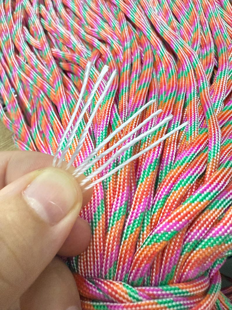 May include: Close-up of a bundle of colorful paracord rope. The rope is a mix of orange, pink, green, and white. A hand holds a few white strands of the rope. The rope is tightly woven and appears durable.