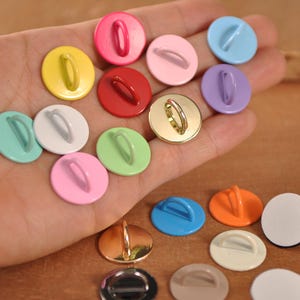 May include: A collection of colorful, round sew-on buttons in various shades including pink, yellow, red, and blue. The buttons have a loop on the back for sewing and are displayed in a hand. Some buttons have a metallic finish.