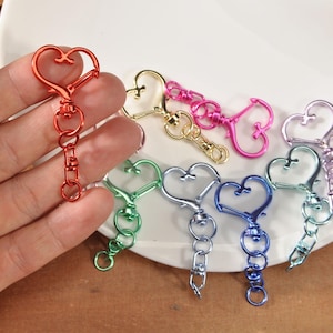 Könnte beinhalten: Eine Sammlung herzförmiger Metall-Schlüsselanhänger in verschiedenen Farben, darunter Rot, Gold, Pink, Grün, Blau und Lila. Jeder Schlüsselanhänger hat einen herzförmigen Verschluss und eine Reihe von Verbindungsringen. Auf weißem Untergrund präsentiert.