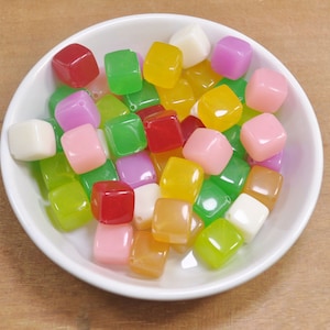 Op de afbeelding: Een kom gevuld met kleurrijke, doorschijnende, vierkante kralen. De kralen zijn in tinten groen, geel, roze, rood en wit.