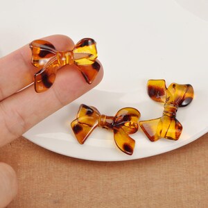 Peut inclure: Trois breloques en forme de nœud papillon de couleur ambre avec un motif écaille de tortue. Les breloques sont faites d'un matériau translucide et sont disposées sur une surface blanche. Les nœuds ont un design classique.