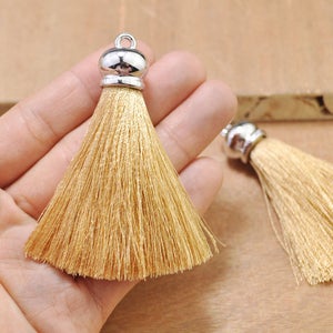 May include: Two beige silk tassels with silver metal caps. The tassels are approximately 2 inches long and are held in a hand.