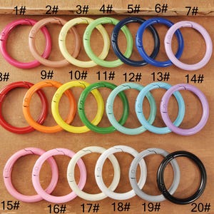 Puede incluir: Una colección de veinte cierres redondos de metal de colores brillantes, incluyendo rosa, naranja, amarillo, verde, azul y negro. Cada cierre tiene una abertura con bisagra para sujetar objetos. Los cierres están numerados del 1 al 20.