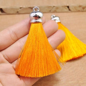 May include: Two bright orange silk tassels with silver metal caps. The tassels are hanging from a hand.