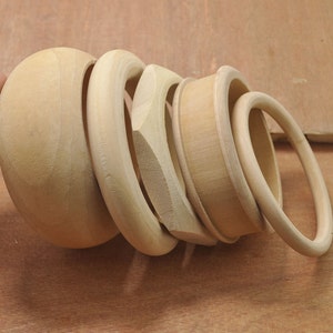 May include: A set of five wooden bangles, all in a light brown color. The bangles are stacked on top of each other, with the largest bangle at the bottom and the smallest bangle at the top.