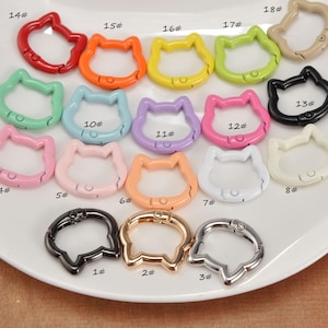 Puede incluir: Una colecci&oacute;n de cierres giratorios con forma de gato en varios colores, incluyendo rojo, naranja, amarillo, verde, rosa, azul, morado, negro, blanco y acabados met&aacute;licos. Estos cierres est&aacute;n dispuestos sobre un plato blanco, mostrando sus diversos tonos y dise&ntilde;os.