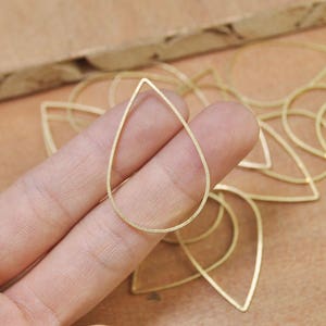 May include: A close-up of a gold teardrop-shaped pendant, with a smooth, polished finish. The pendant is held in a hand, and there are several other similar pendants in the background.