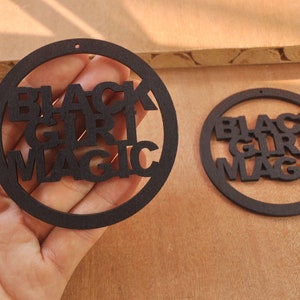 May include: Two black wooden round ornaments with the text "Black Girl Magic" cut out in the center.