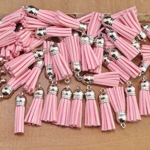 May include: A collection of pink suede tassels with silver metal caps. The tassels are arranged in a random pattern on a wooden surface.