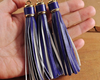 May include: Three purple and white leather tassels with gold metal tops. The tassels are hanging from a hand.
