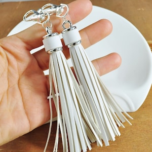 May include: Two white leather tassels with silver metal rings and caps. The tassels are hanging from a silver chain.