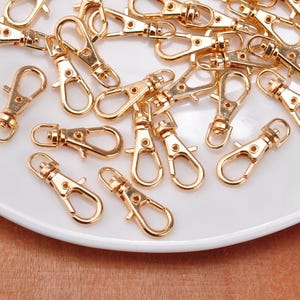 May include: A collection of gold-toned metal lobster claw clasps, each featuring a spring-loaded gate and a swivel base. These small hardware components are designed for attaching items and are scattered across a smooth, white surface.