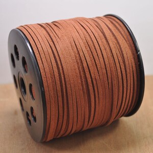 May include: A spool of brown suede cord, wound around a black plastic bobbin. The cord is approximately 3 mm wide.