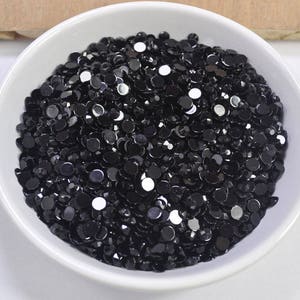 Pode incluir: Uma tigela branca cheia de strass redondos pretos. Os strass são pequenos e têm um acabamento brilhante.