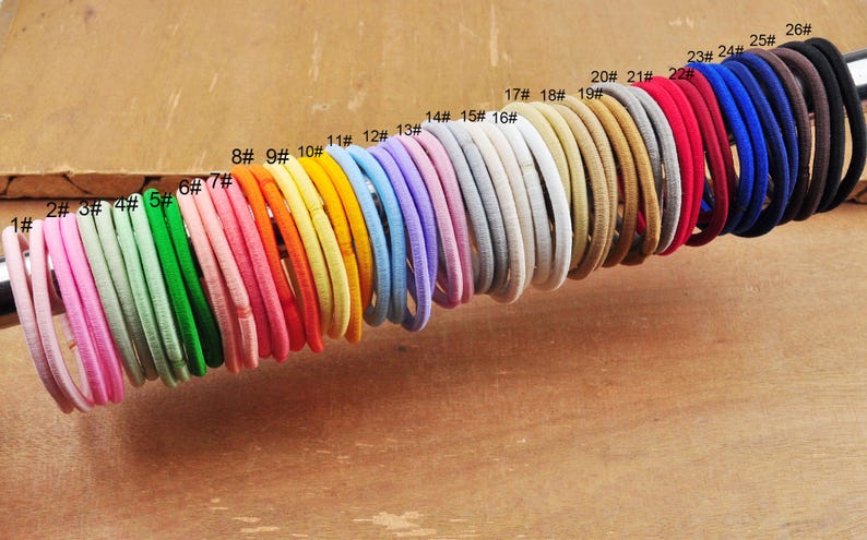 K&ouml;nnte beinhalten: Ein Set mit 26 bunten Haargummis, von 1 bis 26 nummeriert, in einer Reihe angeordnet. Die Haargummis sind aus einem weichen, dehnbaren Material gefertigt und in verschiedenen Farben erh&auml;ltlich, darunter rosa, gr&uuml;n, blau, gelb, orange, rot, braun und schwarz.
