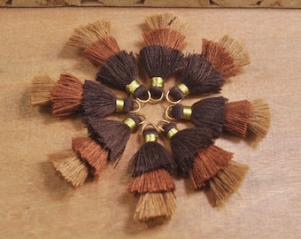 May include: A set of nine brown, tan, and dark brown tassel trims with gold rings. The tassels are arranged in a circle on a wooden surface.