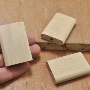 Op de afbeelding: Drie onafgewerkte houten blokken, twee zijn rechthoekig en één is iets langer en heeft een klein gat in het midden.