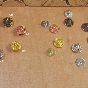 May include: Assortment of 6 different sizes of metal pin backs. The pin backs are numbered 1 through 6. The pin backs are silver, gold, and bronze.
