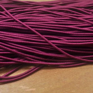 May include: Close-up of a bundle of burgundy elastic cord. The cords are tightly packed together, creating a textured surface. The color is a deep, rich shade of red-purple, with a slight sheen. The background is a light brown wooden surface.